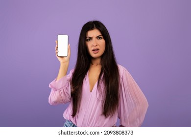 Serious Armenian Lady Holding Smartphone With White Blank Screen, Showing App Or Website, Being Disappointed, Standing Over Purple Background, Mockup