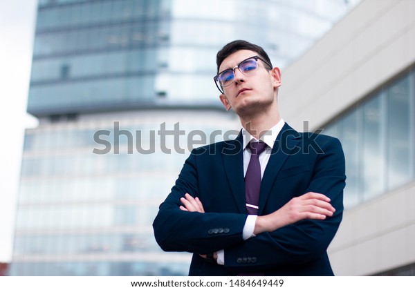 Serious arrogant young businessman in a suit\
with tie and in glasses looking down at camera and holding his\
hands or arms crossed standing outdoor in front of modern office\
business centre,\
skyscraper