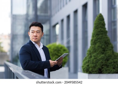 Serious Asian Boss Reads News From Tablet Near Modern Office Male Businessman In Business Suit