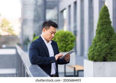 Serious Asian Boss Reads News From Tablet Near Modern Office Male Businessman In Business Suit