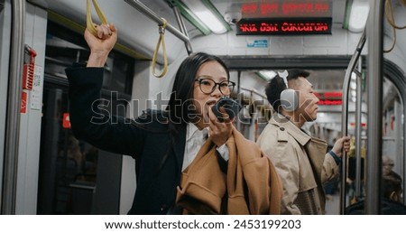 Serious Asian businesswoman holding on to safety handle with one arm. Recording voice message to someone with instructions. People in background standing still and listening to music in headphones.