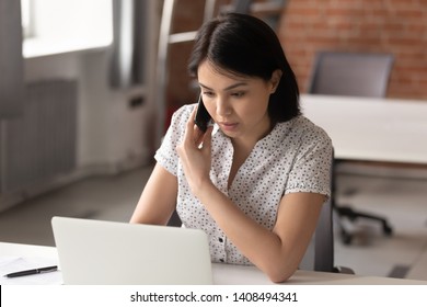 Serious asian businesswoman using laptop looking at computer talking on phone consult client sit at desk, focused female sales manager make mobile business call discuss work with customer in office