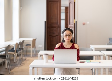 Serious Asian College Girl In Headphones Watching Video Lesson Or Webinar Via Internet On Laptop While Sitting In Empty Reading Room, Female Student Working On Graduation Project In Library