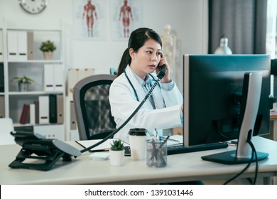 Serious Asian Female Doctor On Phone In Office. Young Japanese Woman Medical Worker In Clinic Hospital Connecting With Nurse On Telephone Call Checking Patient Situation Of Surgery On Computer Screen