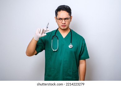 Serious Asian Male Doctor Wearing Rubber Gloves And Scrubs, Looking Amazed At Syringe With Vaccine, Isolated Over White Background