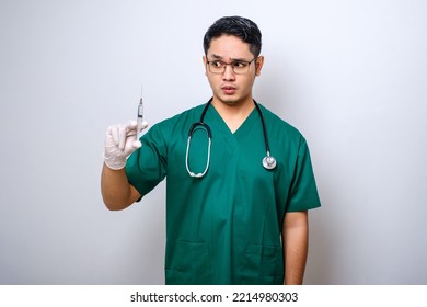 Serious Asian Male Doctor Wearing Rubber Gloves And Scrubs, Looking Amazed At Syringe With Vaccine, Isolated Over White Background