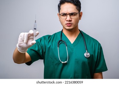 Serious Asian Male Doctor Wearing Rubber Gloves And Scrubs, Looking Amazed At Syringe With Vaccine, Isolated Over White Background