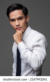 Serious Asian male entrepreneur in white shirt with tie touching chin on gray isolated background in light studio
