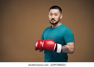 Serious asian man in green t shirt wearing red boxing gloves standing in fight pose and looking to camera. Isolated over brown wall background  - Powered by Shutterstock