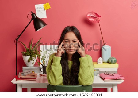 Serious Asian woman keeps index fingers on temples, thinks information over, has long dark hair, poses against coworking space, dressed in casual outfit, feels exhausted during exam preparation