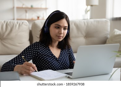 Serious Asian woman wearing headphones looking at laptop screen, writing notes, learning language, focused female student watching webinar, listening to lecture, studying online at home - Powered by Shutterstock