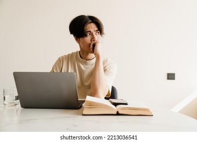 Serious Asian Young Man Studying With Book And Laptop Computer In Bright Room At Home