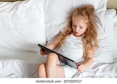 Serious attentive Caucasian Girl use Tablet for watching videos and cartoons. Beautiful Child girl Portrait using tablet Computer lying in bed. Little blond girl play game on tablet in bedroom at home - Powered by Shutterstock