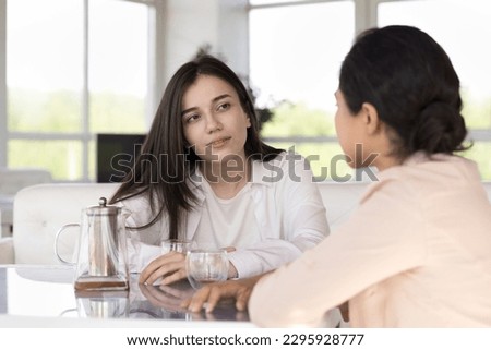 Serious attentive woman spend time with Indian female friend listen her, sit together at table with teapot, having reliable talk meet in cafe. Friendship, relations, honest trustworthy conversation