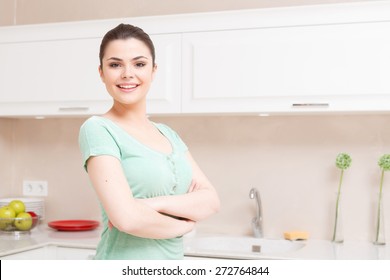 Serious attitude. Young smiling lady standing in kitchen with her arms folded across her chest