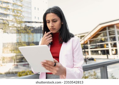 Serious attractive Indian woman holding digital tablet, check email, working online standing outdoors. Pensive asian student studying, learning language in university campus, education concept 