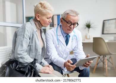 Serious Attractive Mature Woman In Casual Outfit Sitting On Chair In Doctors Office And Listening To Doctor Using Tablet To Analyze Test Results During Appointment