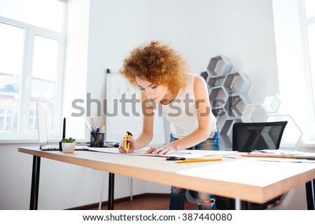 Serious attractive young woman photographer with curly red hair working in office using stationery knife
