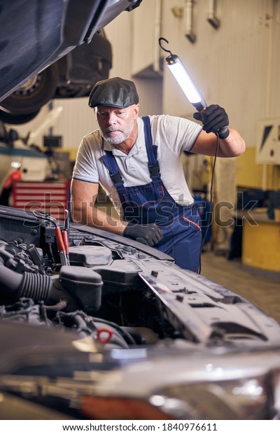 Serious auto\
mechanic in work overalls using LED inspection lamp while repairing\
car in automobile repair\
garage