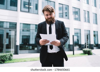 Serious bearded businessman in formal clothes standing on street near modern office building and working with documents during daylight outdoors - Shutterstock ID 1784650661