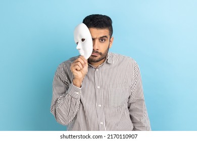 Serious Bearded Businessman Removing White Mask From Face Showing His Strict Expression, Pretending To Be Another Person, Wearing Striped Shirt. Indoor Studio Shot Isolated On Blue Background.