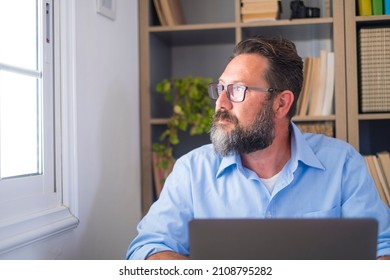 Serious Bearded Caucasian Man In Deep Thoughts Looking Out Through Office Window. Businessman Taking A Break From Work And Admiring View From Workplace. Male Executive Looking Away While Working 