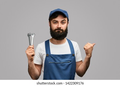 Serious Bearded Engineer With Wrenches Looking At Camera And Pointing Aside With Thumb Against Gray Background