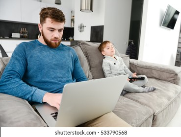 Serious bearded father dressed in blue sweater using laptop while his son watching TV.