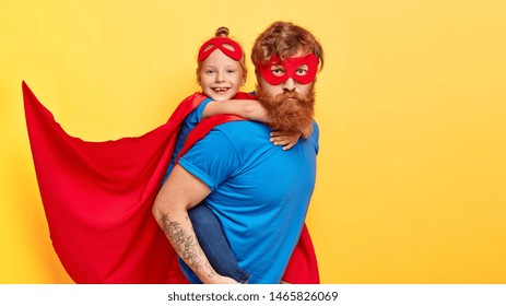 Serious Bearded Ginger Father Superhero Carries Small Female Kid On Back, Do Good Things Together, Wear Costumes With Cape, Ready For Children Party, Isolated On Yellow Wall. We Are Strong Heroes