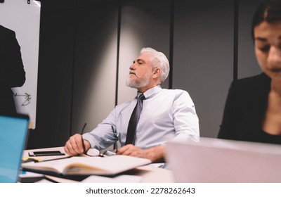 Serious bearded gray haired male executive in classy outfit sitting at table during business conference with colleagues working together on project - Shutterstock ID 2278262643