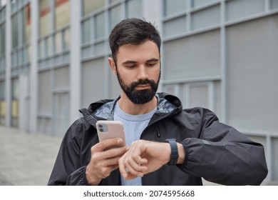 Serious bearded man checks time on smartwatch uses application to monitor fitness activity dressed in black anorak poses outdoors against blurred background. Peope sport and technology concept