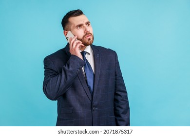 Serious Bearded Man Wearing Dark Official Style Suit Holding Phone Near His Ear, Talks To His Companies, Solving Important Work Issues. Indoor Studio Shot Isolated On Blue Background.