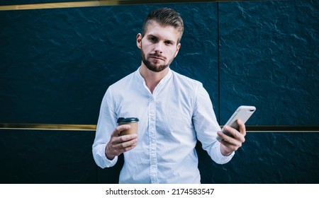 Serious Bearded Man In White Shirt Looking At Camera And Browsing Smartphone While Browsing Smartphone And Holding In Hand Paper Cup With Coffee On City Street. Sports Betting And Earnings 