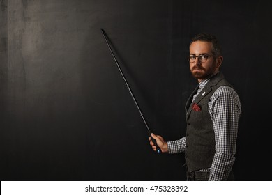 Serious Bearded Professor In Plaid Shirt And Tweed Vest, Wearing Glasses And Looking Condemn On Camera, Shows Something On School Black Board With His Pointer