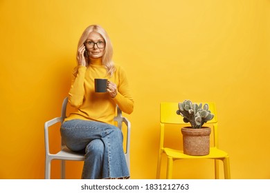 Serious beautiful blonde woman has telephone conversation during coffee break looks directly at camera dressed in casual outfit poses near chair with potted cactus isolated on yellow background