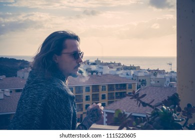 serious beautiful man model stay outside in the terrace at home with nice view of houses and city and ocean horizon. sunset time in summer night. smoking and stay alone concept - Powered by Shutterstock