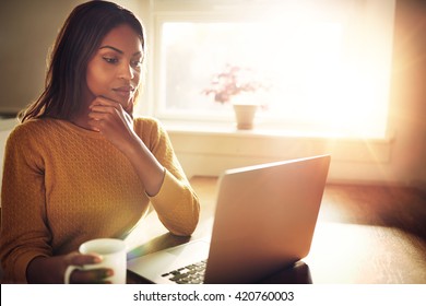 Serious beautiful woman with hand on chin sitting near bright window while looking at open laptop computer on table and holding white mug - Powered by Shutterstock