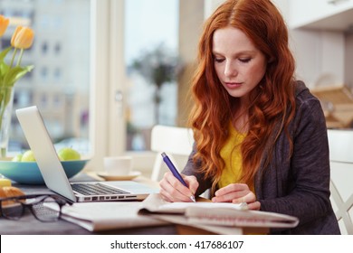 Serious Beautiful Young Adult Woman With Red Hair Taking Notes While Sitting In Front Of Laptop Computer On Kitchen Table Near Window