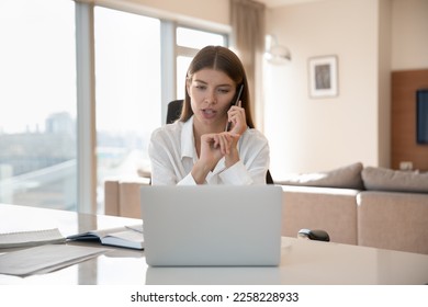 Serious beautiful young freelance professional girl talking to customer on cellphone at home office workplace, giving telephone support, looking at laptop, speaking at table with pc - Powered by Shutterstock