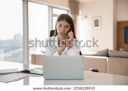 Serious beautiful young freelance professional girl talking to customer on cellphone at home office workplace, giving telephone support, looking at laptop, speaking at table with pc