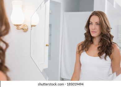 Serious beautiful young woman looking at herself in the bathroom mirror at home