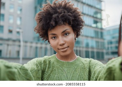 Serious Beautiful Young Woman Stretches Arms And Makes Selfie During Walk In Urban Setting Wears Casual Green Jumper Has Confident Look At Camera. Female Blogger Records Video To Front Camera