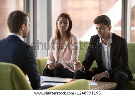 Serious biracial businesswoman and middle eastern ethnicity business partners listening caucasian businessman sitting in comfortable couches negotiating discussing startup new project in modern office