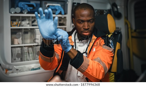Serious Black African American Paramedic Puts on\
Blue Surgical Rubber Gloves in an Ambulance Vehicle with an Injured\
Patient. Emergency Medical Technician is Checking Up on Victim\'s\
Vitals.