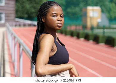 Serious black female with long braids stands leaning on metal fencing looking at camera. Portrait of African American woman standing on sports ground of residential complex on blurred background - Powered by Shutterstock