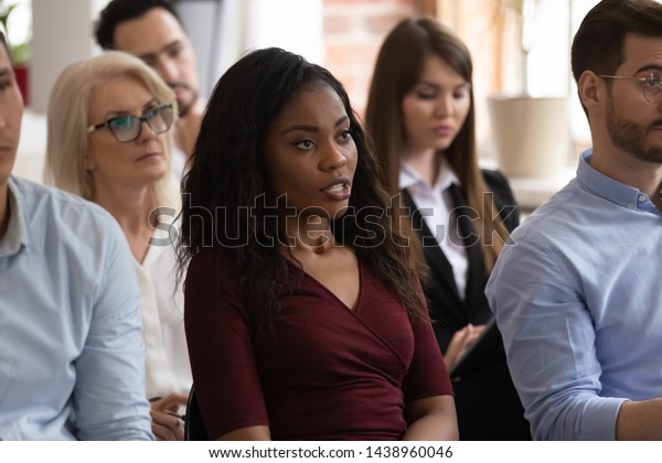Serious black female professional ask question\
at group seminar lecture event, confident african business woman\
training conference participant sit on chair talking at corporate\
meeting workshop