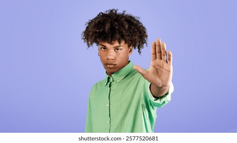 Serious black guy gesturing STOP, saying NO, showing rejection gesture, expressing refusal or denial, standing on blue studio background. Negative opinion concept