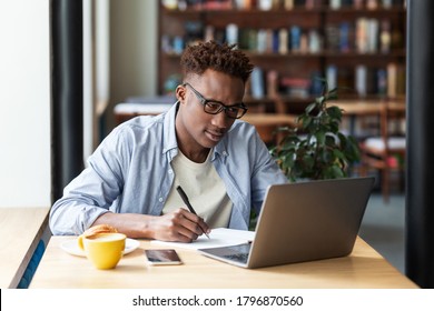 Serious Black Guy Taking Notes While Studying Online In Front Of Laptop At City Cafe