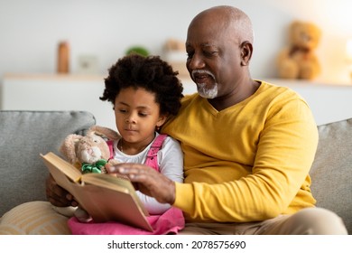 Serious Black Small Girl With Toy And Elderly Man Reading Book With Fairy Tales On Sofa In Living Room Interior. Free Time Together, Relationship Of Grandfather And Granddaughter, Love And Family