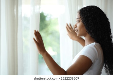 Serious black woman opening curtains and looking through a window at home - Powered by Shutterstock